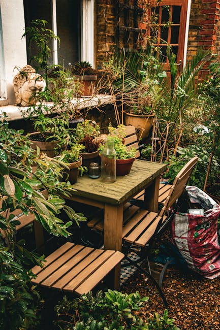 A cozy backyard garden patio featuring a weathered wooden table surrounded by matching wooden chairs and a metal-framed chair, all set on a small gravel surface. The table is decorated with an assortm